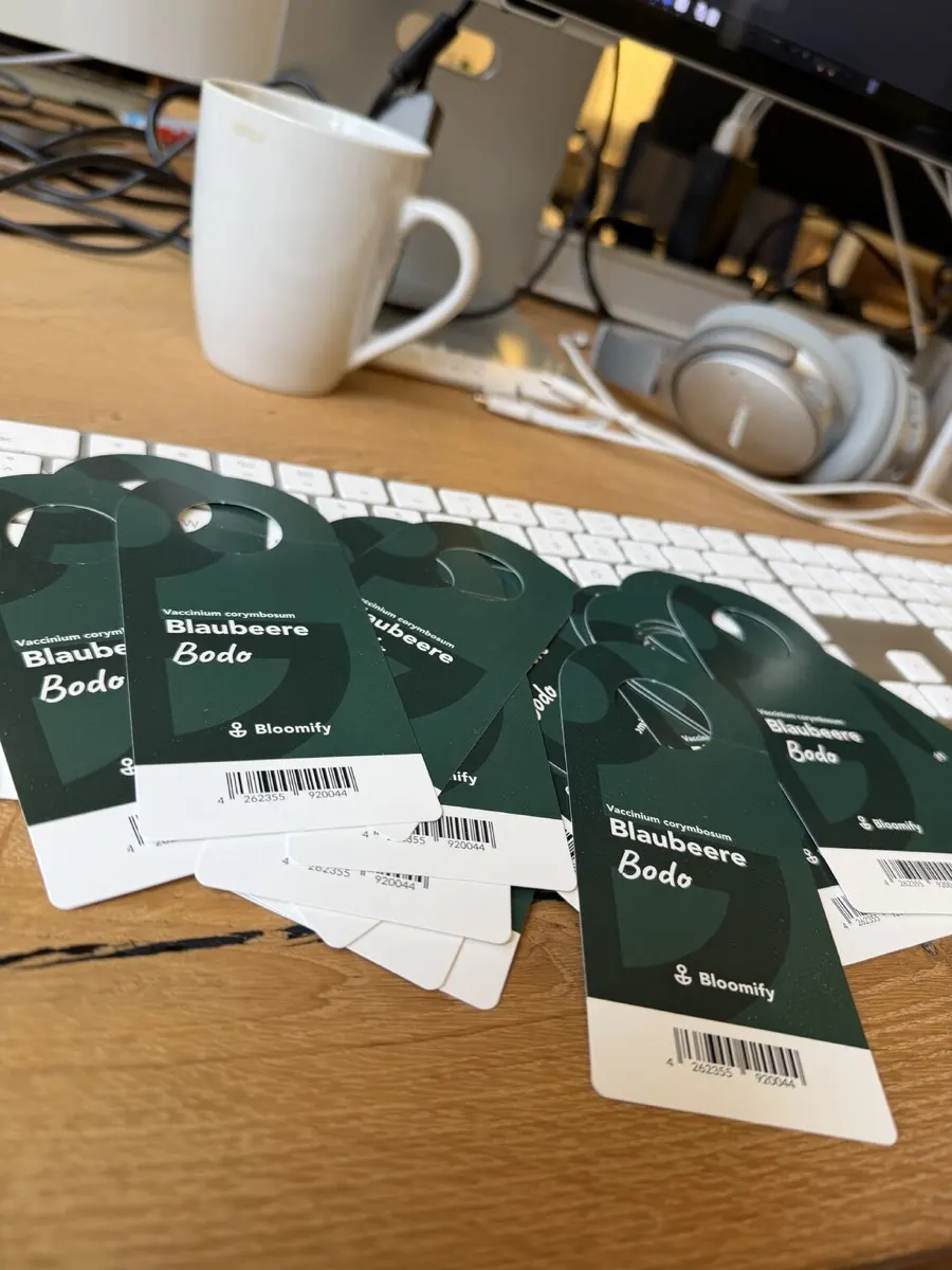 Gedruckte Pflanzen-Einstecker „Blaubeere Bodo“ von Bloomify mit Barcode auf einem Schreibtisch.